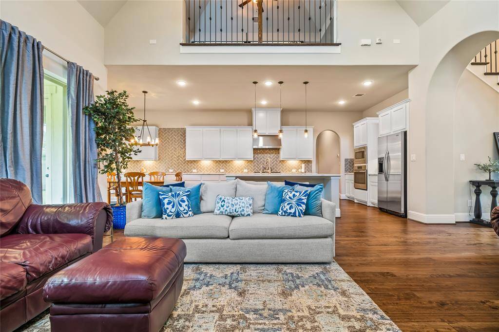 This inviting open-concept living space features a cozy seating area with a plush sofa and accent pillows, leading into a modern kitchen with white cabinetry, stainless steel appliances, and stylish pendant lighting. The warm hardwood floors and high ceilings enhance the spacious feel, while large windows provide ample natural light.