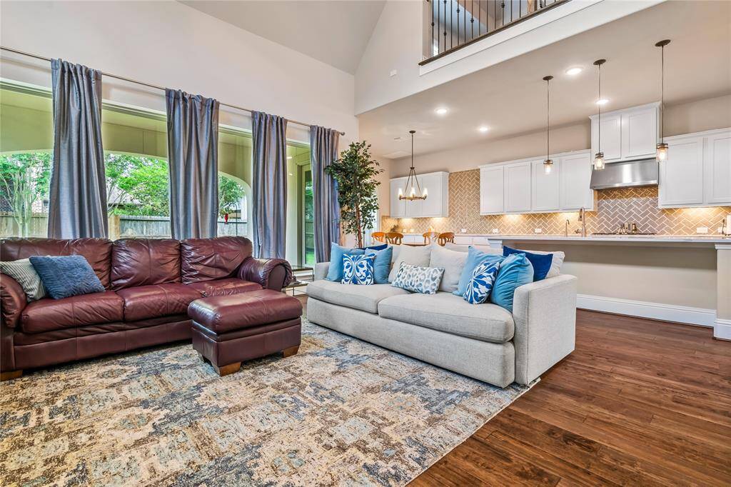 This spacious living area features a high ceiling and an open floor plan, combining a cozy seating area with a modern kitchen. Large windows provide abundant natural light, overlooking a lush backyard. The warm wood flooring complements the stylish decor, creating an inviting atmosphere.