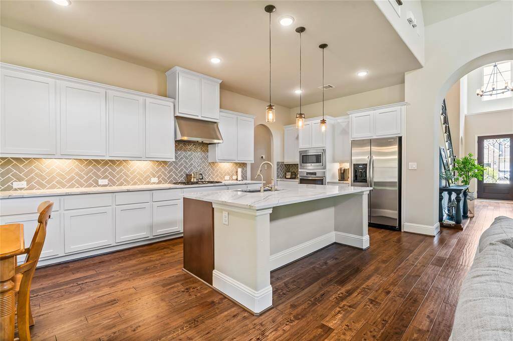 This spacious kitchen features elegant white cabinetry, a large island with a marble countertop, and stainless steel appliances. The herringbone backsplash adds a stylish touch, while pendant lighting and wood flooring create a warm, inviting atmosphere.