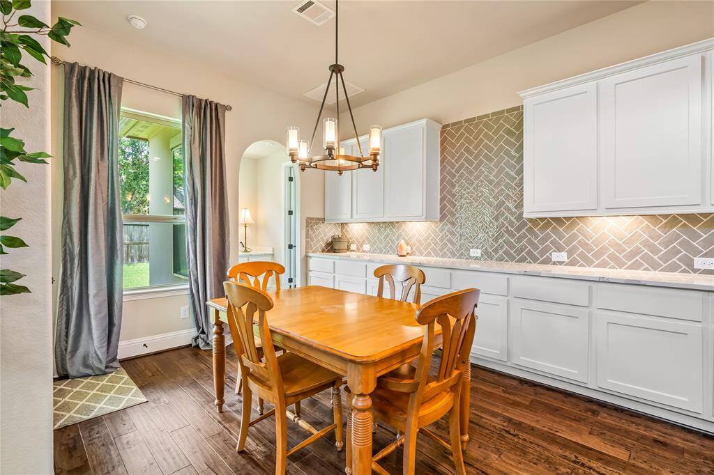 Bright and inviting, this breakfast area features a wooden table for four, elegant lighting, and ample white cabinetry. Enjoy views of the lush outdoors through a large window, complemented by a stylish herringbone backsplash.