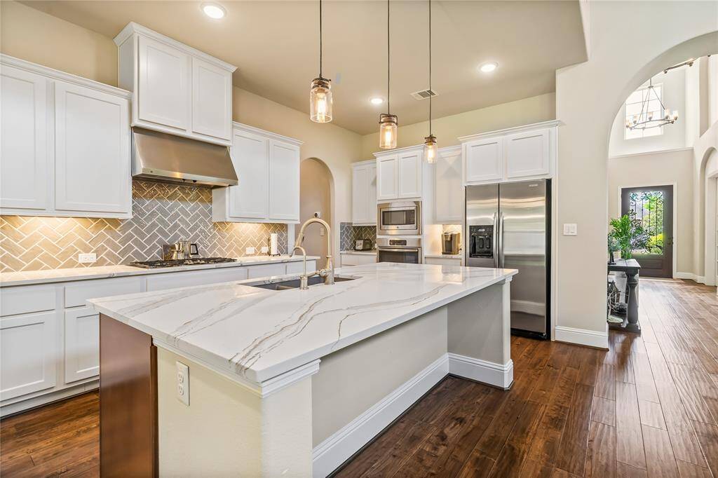 Bright and spacious kitchen featuring white cabinetry, a large Quartzite island with pendant lighting, stainless steel appliances, and elegant herringbone backsplash. Warm wood flooring adds a cozy touch.