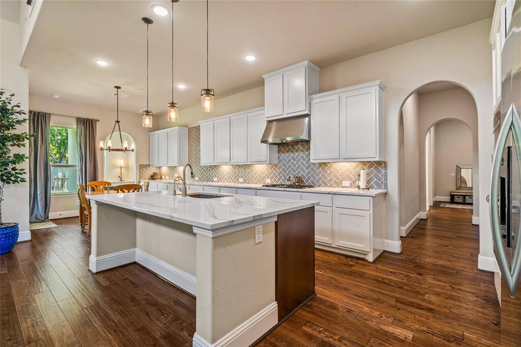 This modern kitchen features a spacious island with a sleek, Quartzite countertop, pendant lighting, and white cabinetry. The open layout flows into a bright dining area with large windows and hardwood floors, creating an inviting atmosphere perfect for entertaining.