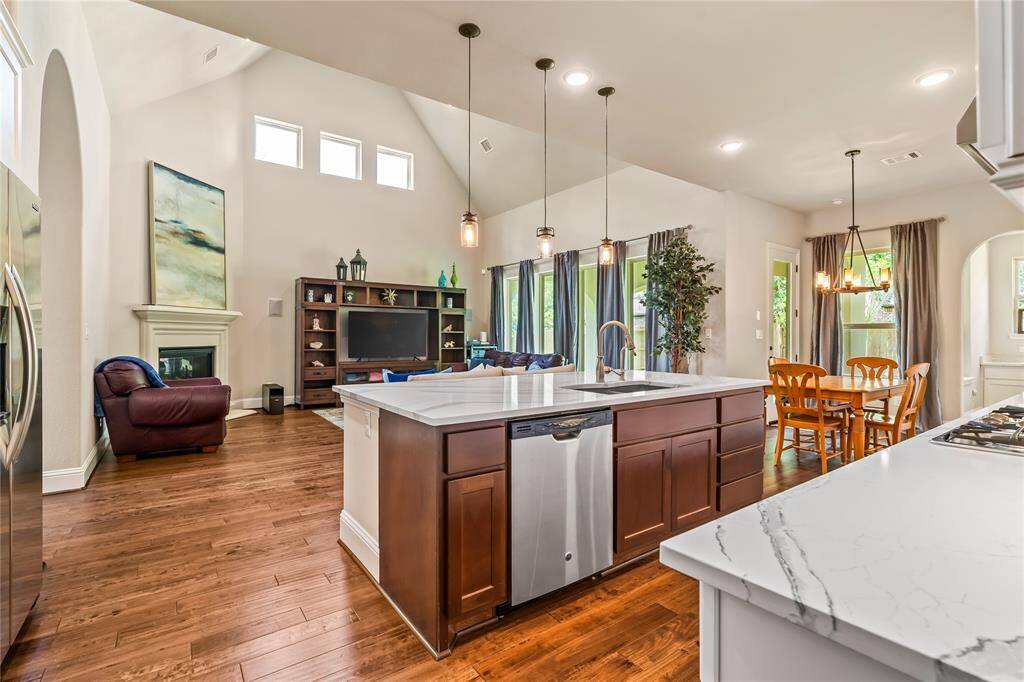 This open-concept living area features high vaulted ceilings, abundant natural light, and hardwood flooring. The spacious, Quartzite kitchen island with modern appliances flows seamlessly into the cozy living room with a fireplace and a dining area, perfect for entertaining.
