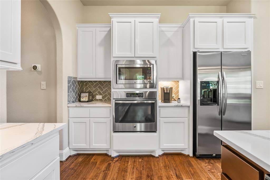 This kitchen features sleek white cabinetry, stainless steel appliances including a double oven and fridge, and a stylish herringbone backsplash. It has hardwood floors and ample counter space(Quartzite) for functionality and style.