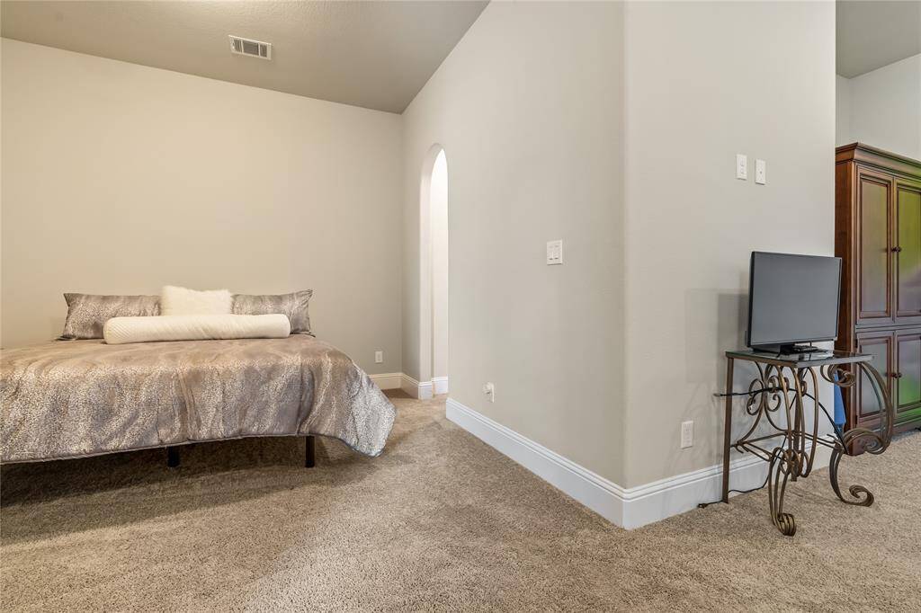 This cozy bedroom features a plush carpet, a comfortable bed with elegant bedding, and a small TV on a decorative stand. The room has soft lighting and an arched doorway leading to another space, creating a warm and inviting atmosphere.