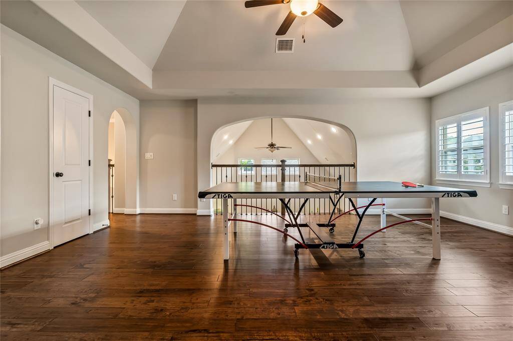 This spacious game room features beautiful hardwood floors, a vaulted ceiling with a fan, and ample natural light from two windows. It includes a ping pong table and opens into a cozy adjoining room, perfect for entertainment.