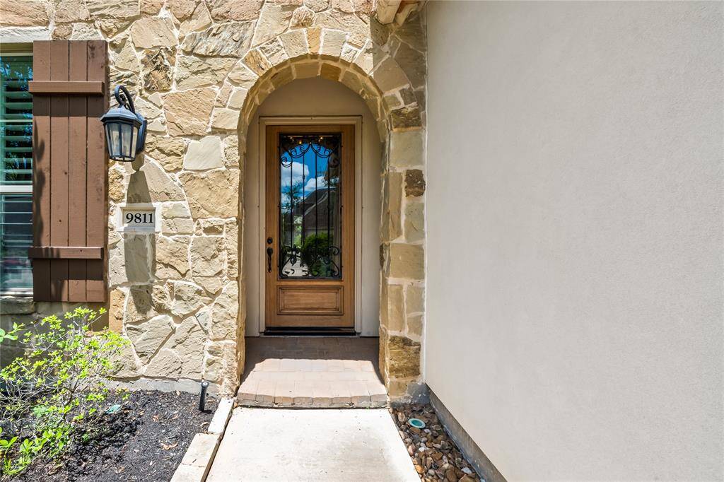 Charming entryway with a stone arch and wooden door, accented by a lantern-style light fixture. The house number is prominently displayed, and there's a small garden area to the side.