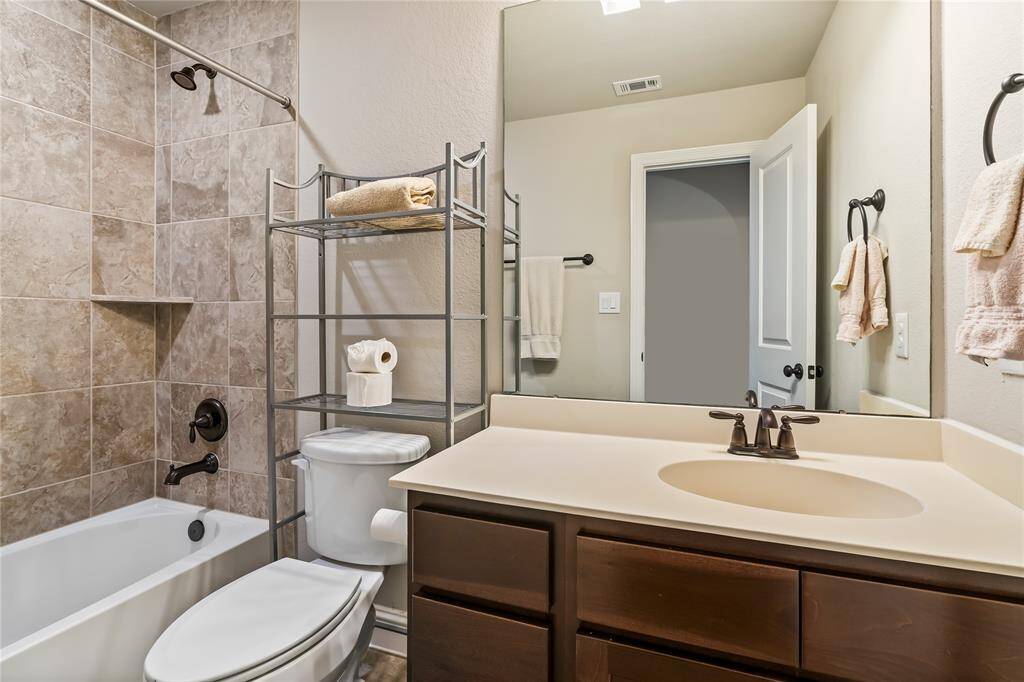 This bathroom features a clean, modern design with a tiled bathtub and shower area, a white vanity with a dark faucet, and neutral-toned walls. It includes a large mirror, ample lighting, and towel racks for convenience.