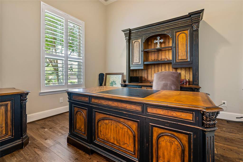 This photo showcases a sophisticated home office with elegant wooden furniture, featuring a large desk and a matching hutch. Natural light filters through a window with plantation shutters, highlighting the hardwood floors and creating a warm, inviting atmosphere.