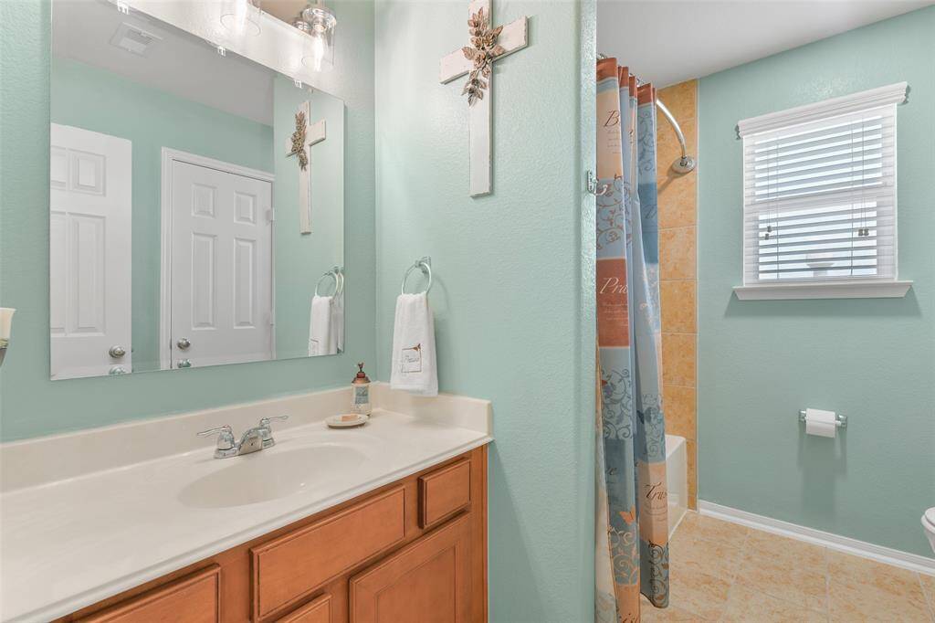 This bathroom services the previous bedroom and the next, featuring a lengthy cultured marble vanity with a single basin sink, wood cabinetry, Edison bulb lighting, tile floors, and a combination bathtub and shower with a tile surround.