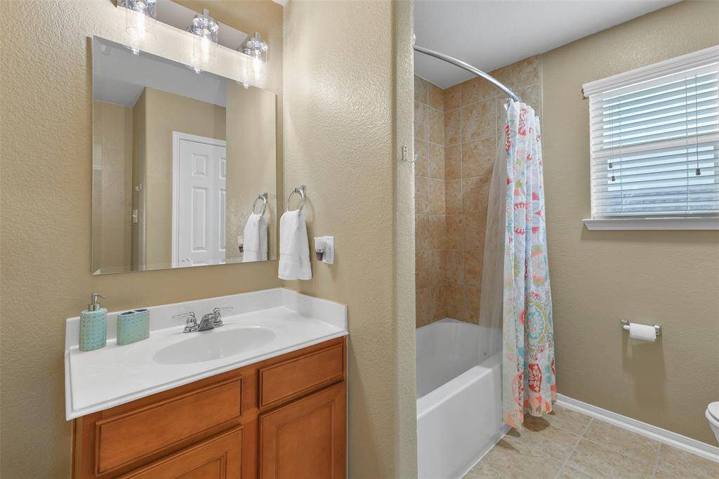 This bathroom services the previous bedroom and it boasts a single cultured marble vanity with a basin sink, wood cabinetry, Edison bulb lighting, tile floors, and a combination bathtub and shower with a tile surround.