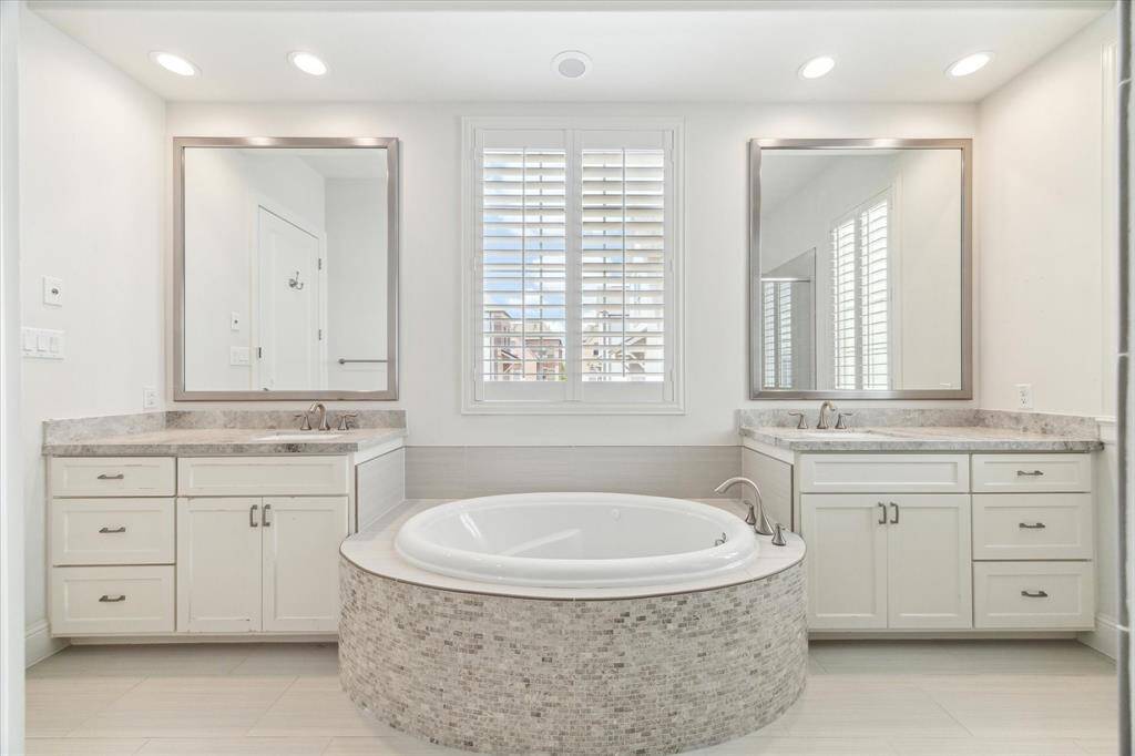 This bathroom features a spacious design with dual vanities, each with a large mirror and ample cabinet storage. A luxurious soaking tub is centrally positioned beneath a window with plantation shutters, offering both elegance and privacy.