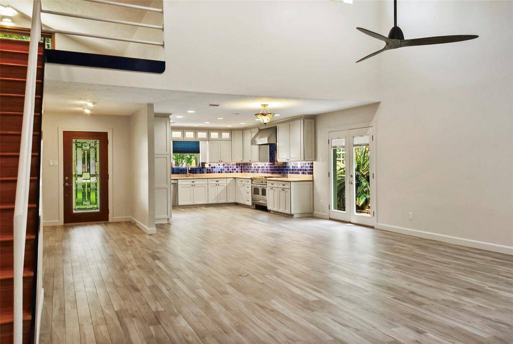 This spacious open-concept living area features light wood flooring, a modern kitchen with a colorful backsplash, and ample natural light from large windows and glass doors. The area includes a stylish staircase and a sleek ceiling fan, creating a bright and airy atmosphere.