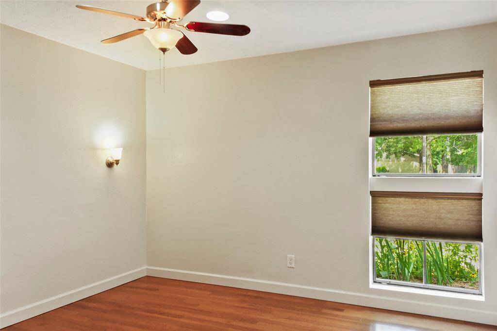 This room features warm wooden flooring and neutral walls. It includes a ceiling fan with lights and two windows with shades, allowing for natural light and garden views.