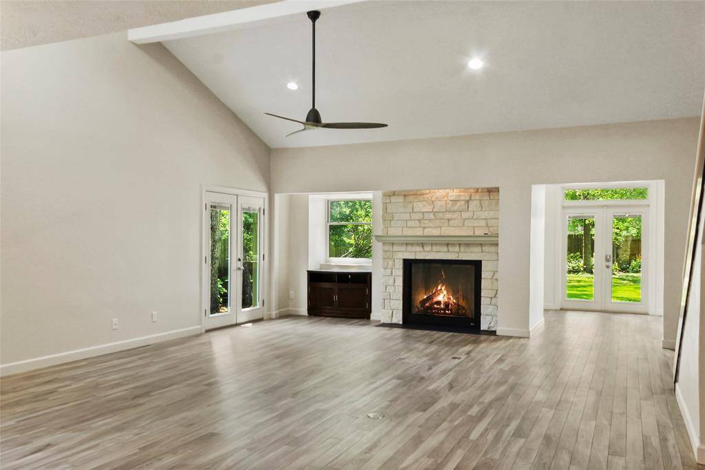 This spacious living room features high ceilings, a modern ceiling fan, and a cozy stone fireplace. Large windows bring in natural light, and a sleek staircase adds a contemporary touch. Perfect for comfortable living and entertaining.