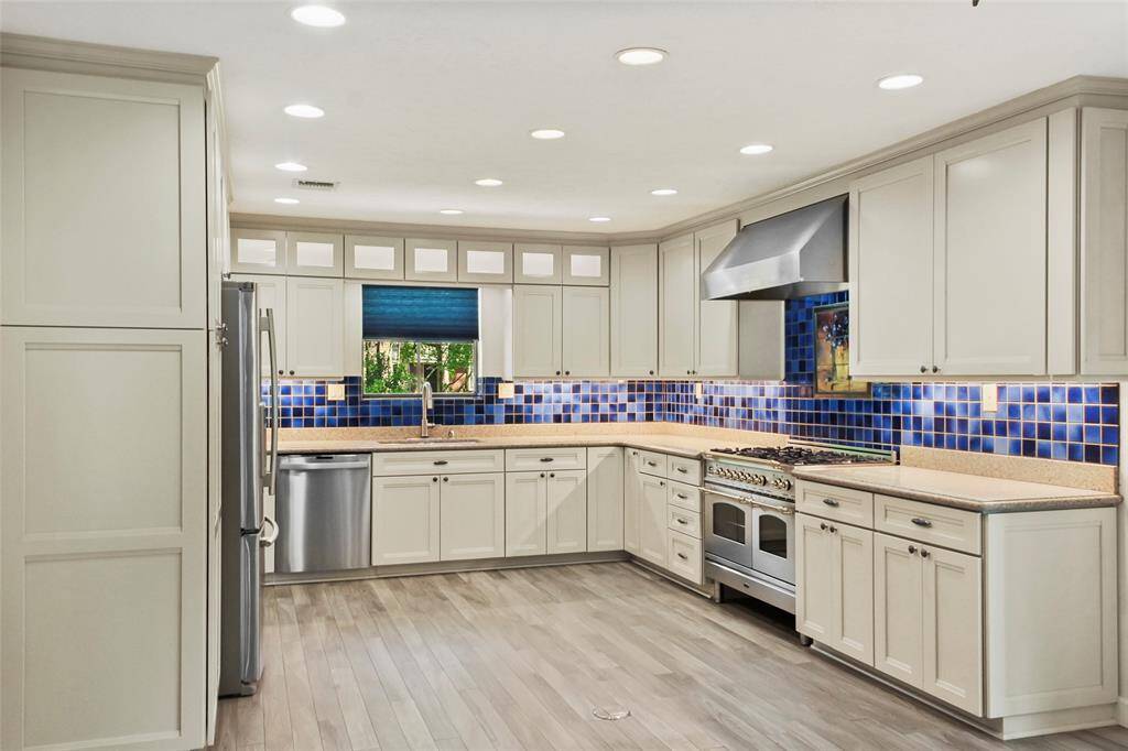 This is a modern kitchen featuring stainless steel appliances, white cabinetry, and a vibrant blue tile backsplash. The room is well-lit with recessed lighting, has ample counter space, and a large window overlooking greenery.