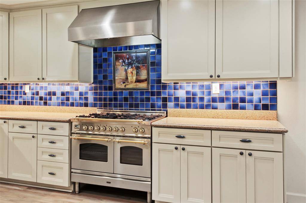 This kitchen features elegant white cabinetry, a stainless steel fancy stove with a large hood, and a striking blue tile backsplash that adds a pop of color.