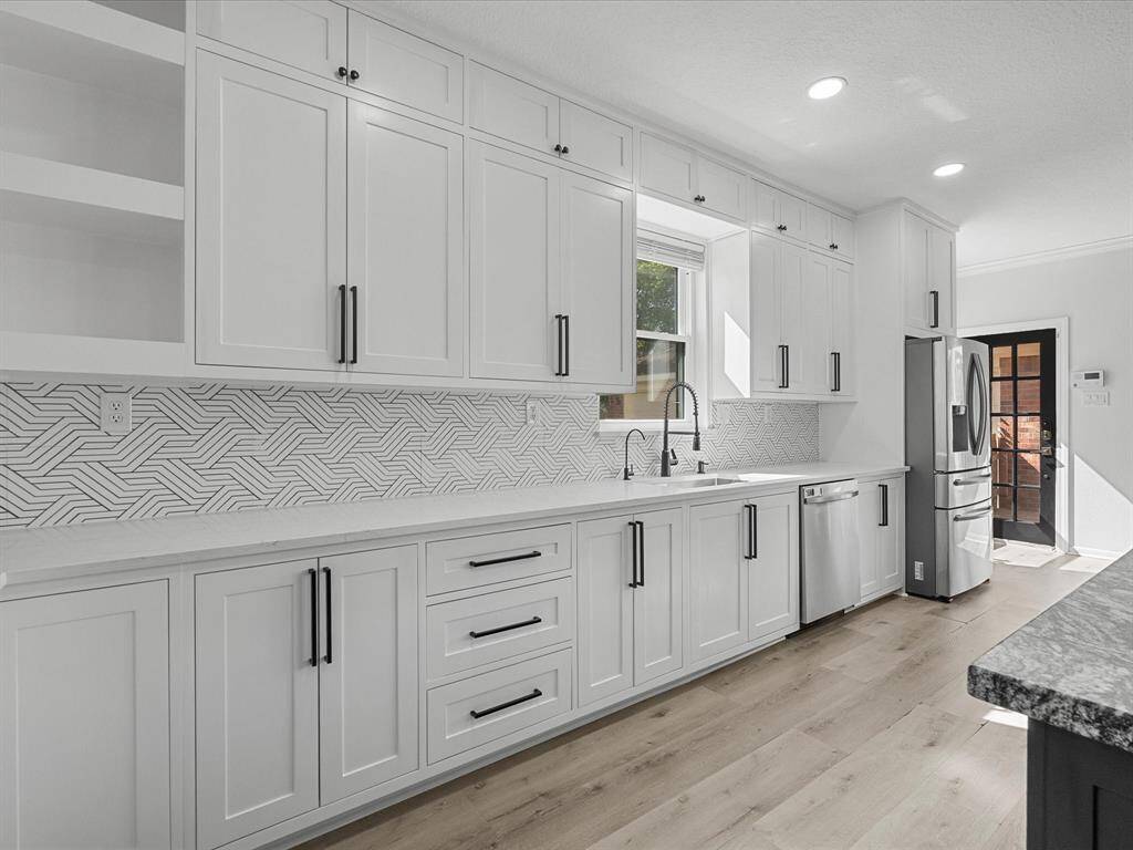 Bright and modern kitchen featuring sleek white cabinetry, elegant geometric backsplash, and stainless steel appliances. The space is enhanced by ample natural light and stylish black hardware, creating a contemporary and inviting atmosphere.