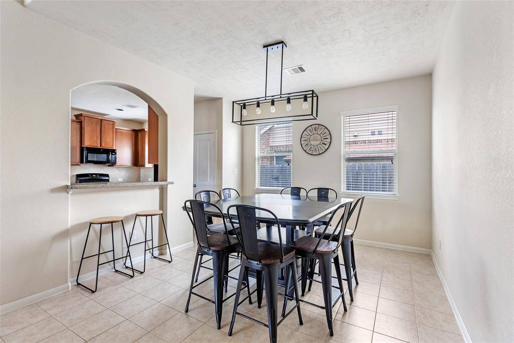 Bright dining area with tile flooring and a modern light fixture. It features an open archway to the kitchen with a bar counter, and windows providing natural light.
