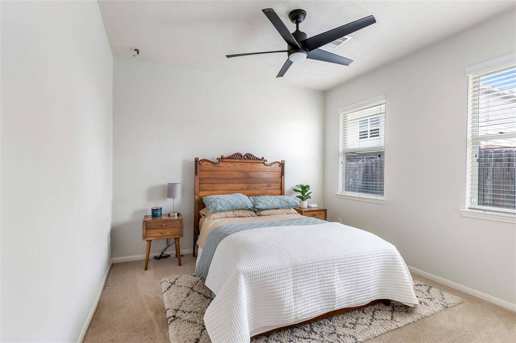 Bright bedroom featuring a cozy setup with soft carpeting, and a ceiling fan. Natural light filters through two windows, enhancing the room's inviting atmosphere.