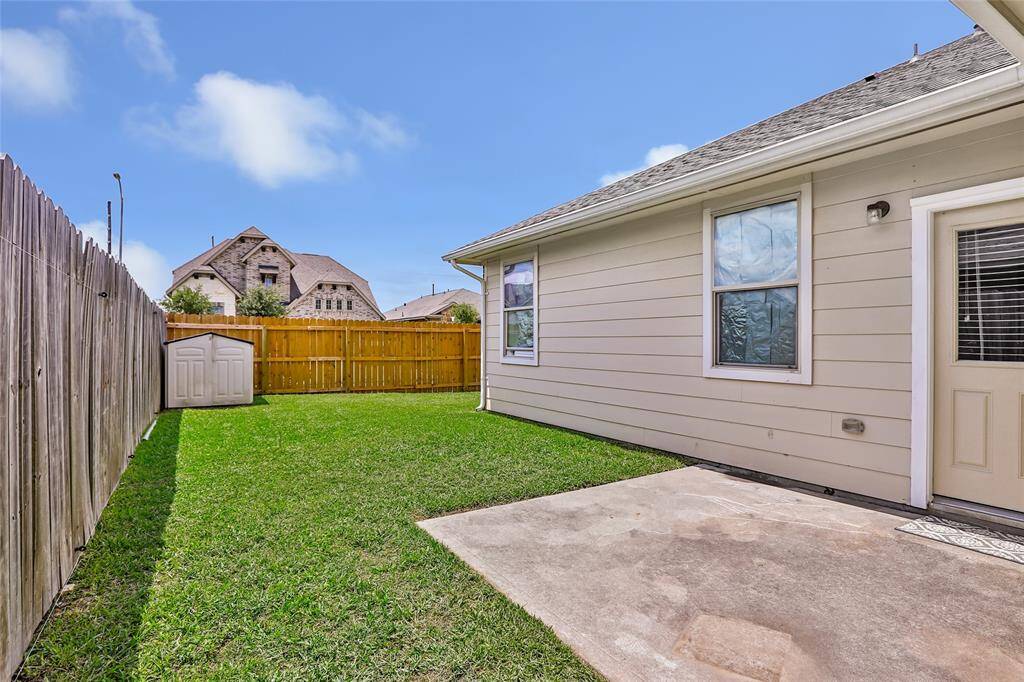 This photo shows a backyard with a well-maintained lawn, a small storage shed, and a concrete patio. The yard is enclosed by a wooden fence, providing privacy. The exterior of the house features light-colored siding and windows.