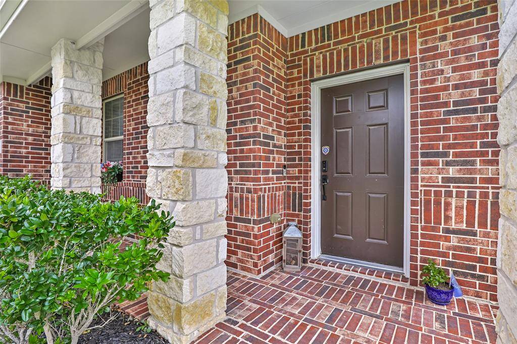 Welcoming front entrance with a sturdy brown door set against a backdrop of red brick walls. It features stone pillars and a small garden with greenery, offering a charming and inviting first impression.