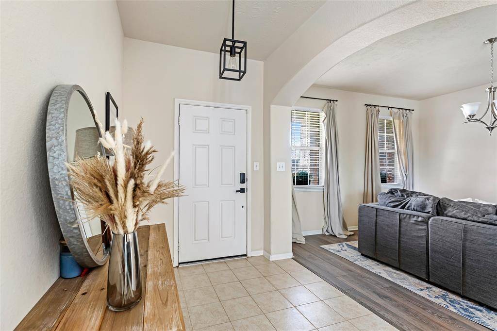 Welcoming entryway with a modern light fixture. The space opens into a cozy living area with natural light from two windows, and a stylish chandelier. The combination of tile and wood flooring adds a warm touch.