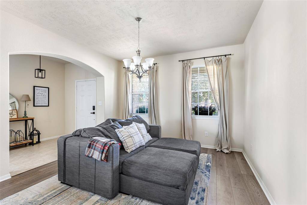 Bright and inviting living room with elegant chandelier, and large windows with curtains. It features a welcoming archway leading to the entrance, complemented by light wood flooring and neutral walls.
