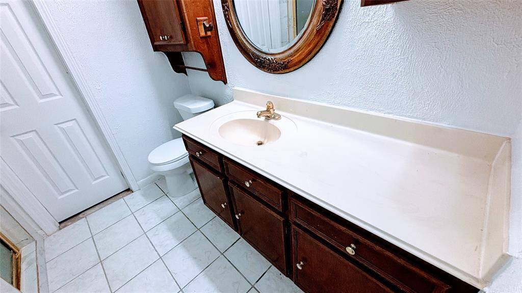 This  bathroom features a single sink vanity with a large countertop, wooden cabinets, and a classic round mirror. It has a tiled floor, a toilet, and a neutral color scheme.