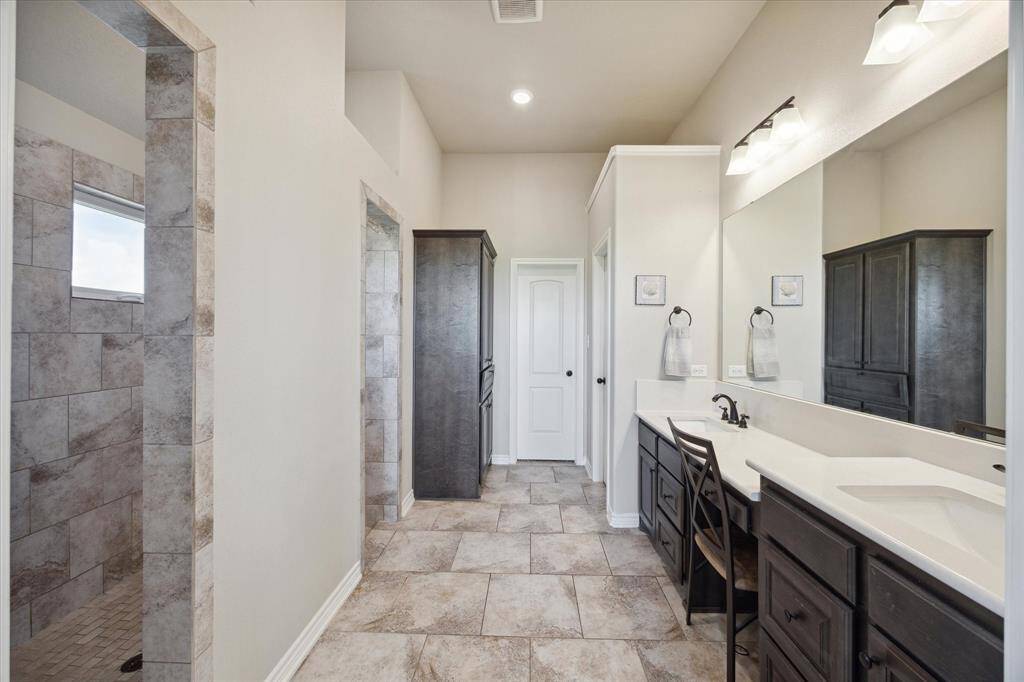 This is a spacious, modern bathroom featuring dual sinks with a large vanity, dark wood cabinets, tiled flooring, and a separate walk-thru shower area visible to the left. It has ample lighting and a neutral color palette.