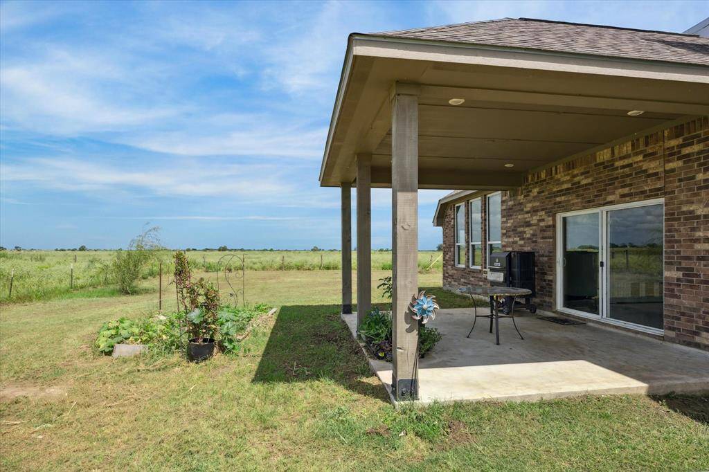 Featuring a covered patio area with a view of an expansive grassy field. The outdoor space includes a small garden plot and room for outdoor dining or recreation.