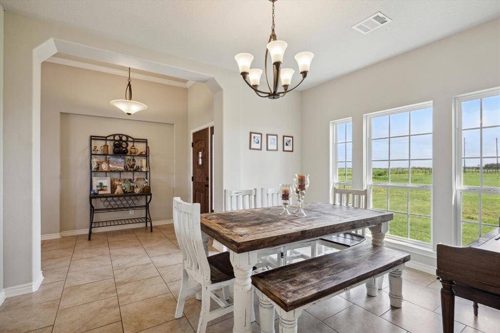 This is a spacious dining area with large windows offering plenty of natural light and a view of green fields. It features an elegant light fixture, with tile flooring and a neutral color palette that creates a warm and inviting space.