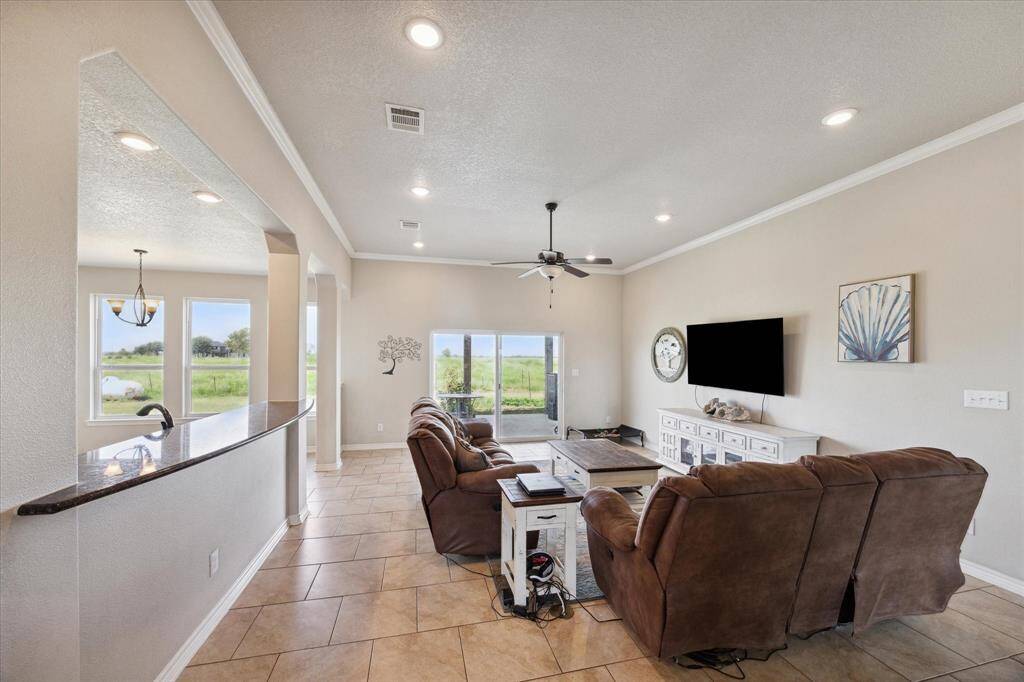 This is a spacious and well-lit living space featuring tiled flooring, a neutral color scheme, and a comfortable seating area. The room offers a scenic view through large windows and includes modern amenities like recessed lighting and a ceiling fan. It seamlessly connects to an adjacent kitchen with a convenient serving bar.