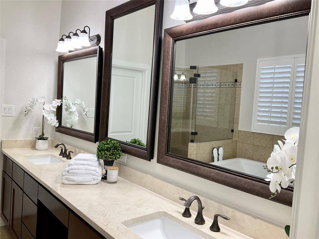 This elegant bathroom features a double vanity with dark wood cabinets and marble countertops. It includes large framed mirrors, modern light fixtures, and a spacious glass-enclosed shower. White shutters provide privacy yet natural light when starting your day.