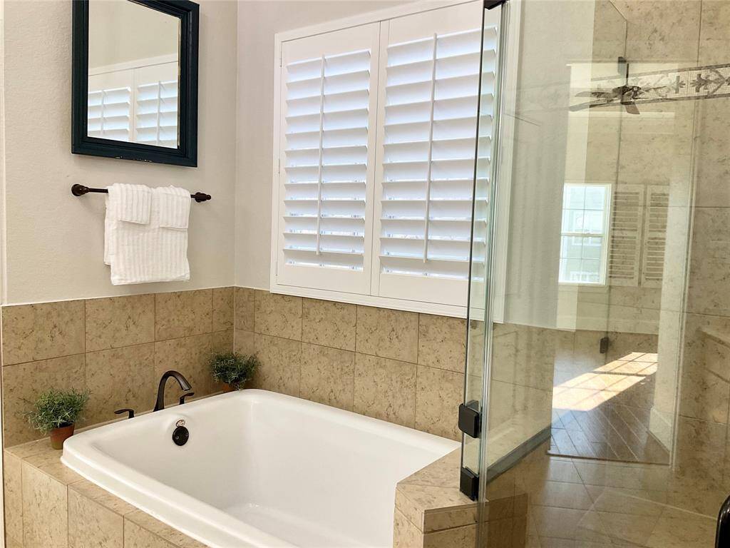 This bathroom features a spacious soaking tub with beige tile surround, a glass-enclosed shower, and a large window with white shutters. A framed mirror and towel rack add practical touches.