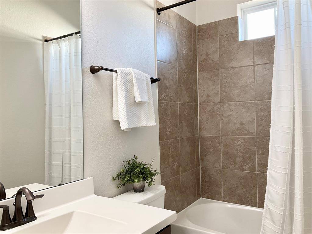 An ensuite bathroom features a clean, modern design with neutral-toned tiles surrounding a bathtub. It includes a white countertop with a sleek faucet, a large mirror, and a small window for natural light. A towel rack and simple decor add a touch of elegance.