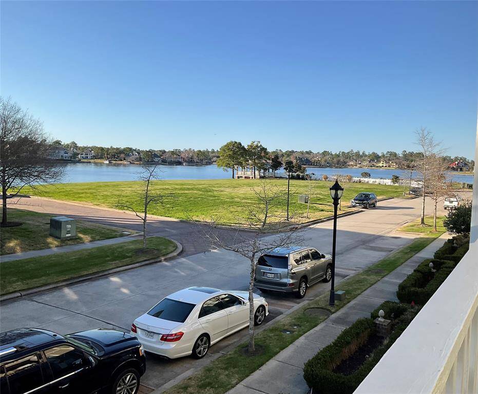 This photo showcases a serene, well-maintained residential area with a lake view. It features a grassy park area, a peaceful street with parked cars for guests only and a picturesque backdrop of Lake Woodlands. Ideal for a tranquil living experience for your family and an active lifestyle.