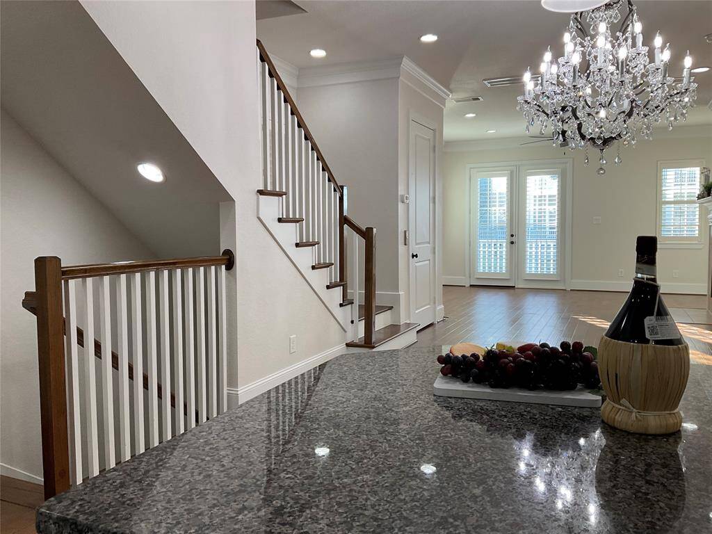 This elegant home interior features a staircase with wooden handrails, an open living space with hardwood floors, and a stunning chandelier. The area is bright with natural light streaming through French doors, and there's a stylish granite countertop ideal for entertaining.