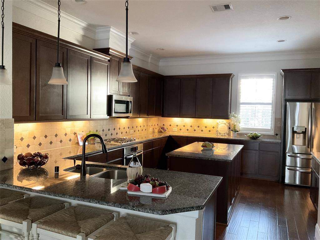 This kitchen features dark wood cabinets, granite countertops, and stainless steel appliances. It includes a central island, pendant lighting, and a tiled backsplash. The space is brightened by natural light from a large window, creating a welcoming and functional cooking area.