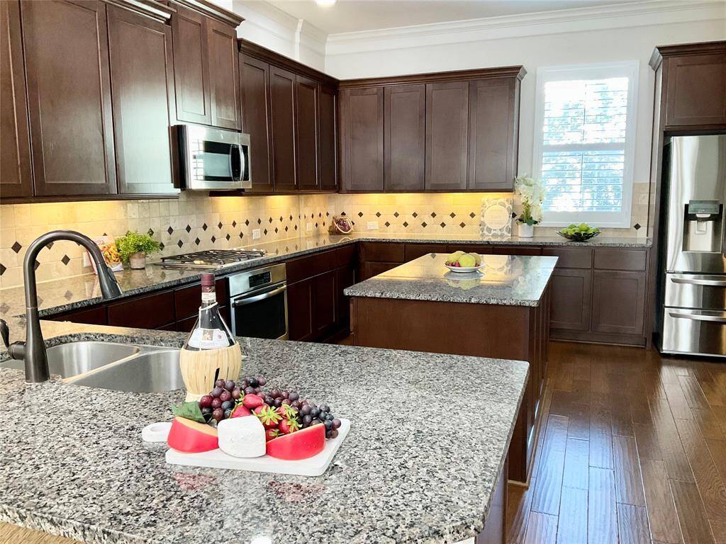 This kitchen features dark wood cabinets, granite countertops, and a central island. Stainless steel appliances, including a built-in microwave and refrigerator, complement the space. The backsplash is tiled with a diamond pattern, and natural light streams in through a window with shutters, creating a warm and inviting atmosphere. Don't miss the large walk in pantry.