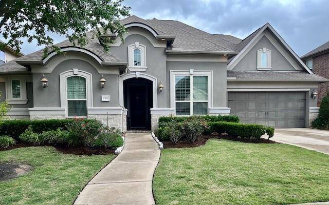 A modern, single-story home with a gray exterior and elegant architectural details. It features a well-maintained lawn, a curved walkway leading to a prominent front door, and a three-car garage with epoxy flooring. The property is surrounded by landscaped shrubs and trees, creating an inviting curb appeal.