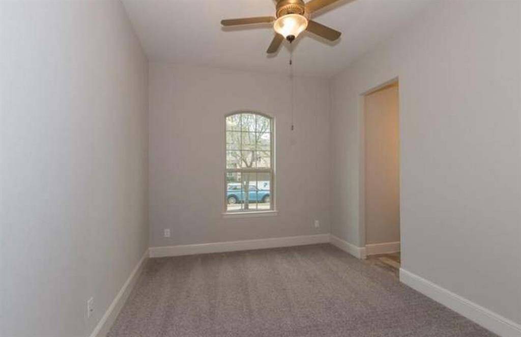 This room features soft gray walls, a ceiling fan with a light, and a carpeted floor. A single arched window allows natural light and offers a view outside. It's compact and versatile, suitable for an office or small bedroom.