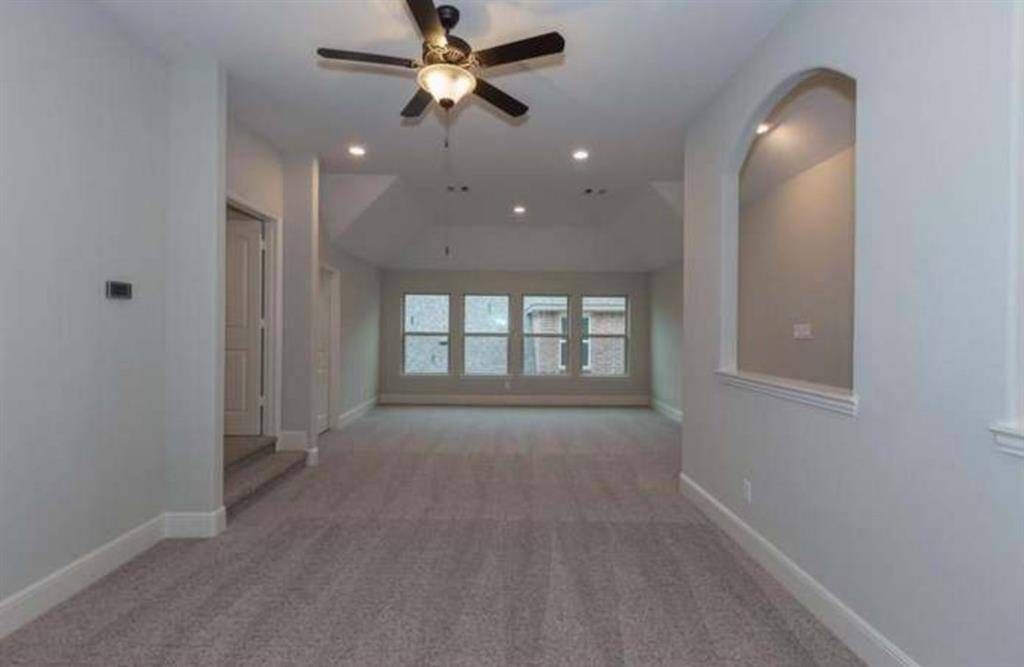 This spacious room features a neutral color palette with plush carpeting, recessed lighting, and a ceiling fan. It has large windows for natural light and an arched wall cutout for added character.