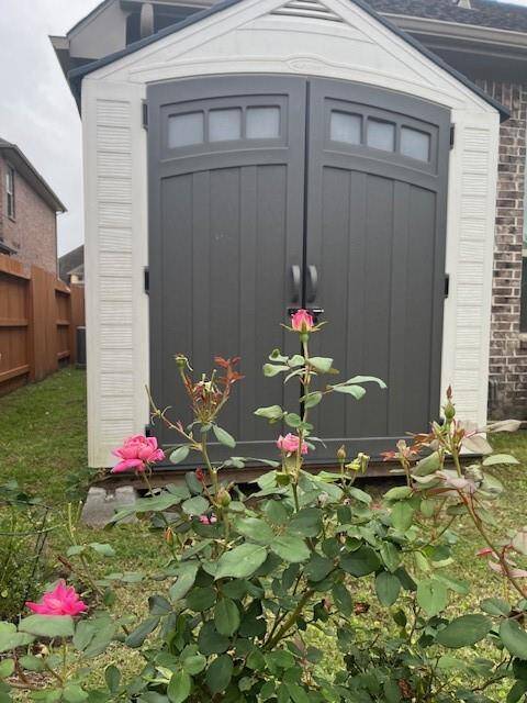 A backyard storage shed with double doors, set against a brick wall. In the foreground, there are blooming pink roses, adding a touch of color to the garden area.
