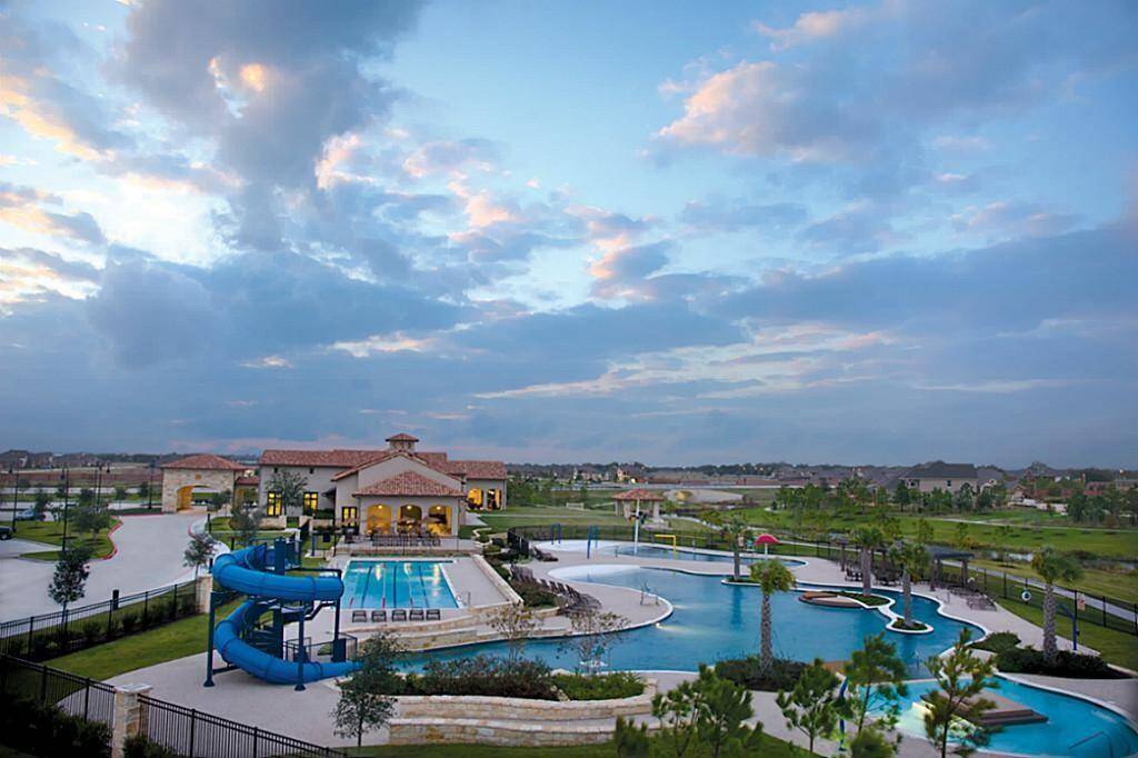 A luxurious community recreation area featuring a large swimming pool with a slide, surrounded by greenery. A clubhouse with a tiled roof and ample windows is visible in the background, offering a scenic view of the neighborhood under a picturesque sky.