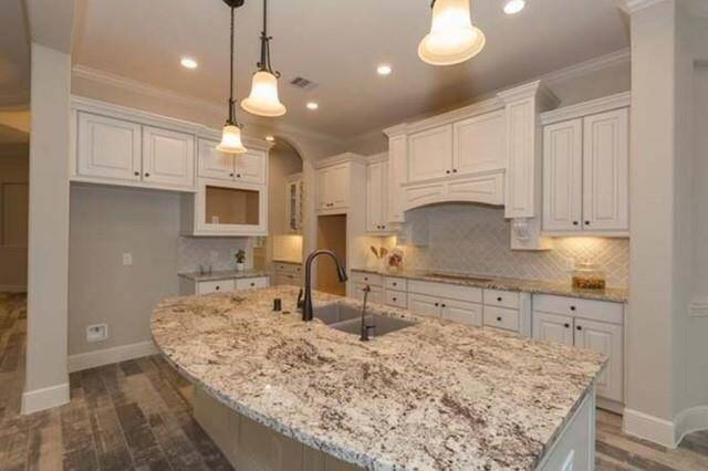 This kitchen features a spacious, curved granite island with a modern dual sink and elegant pendant lighting. It boasts white cabinetry, a neutral backsplash, and recessed lighting, creating a bright and inviting space. Perfect for cooking and entertaining.