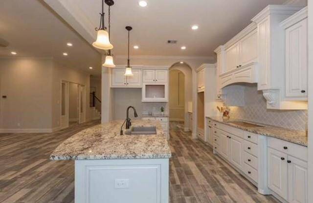 A spacious, modern kitchen featuring granite countertops, a large island with a sink, elegant pendant lighting, and ample white cabinetry. The open layout and wood-look tile flooring add to its appeal, offering both functionality and style.