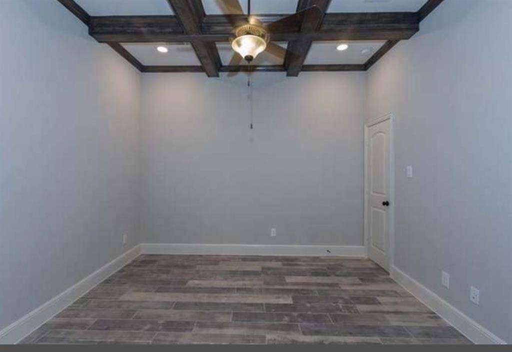 This room features a modern design with light gray walls, wood-look tile flooring, and a ceiling fan. The coffered ceiling with dark wood beams adds character, and a single door provides access. Ideal for a cozy study or small bedroom.