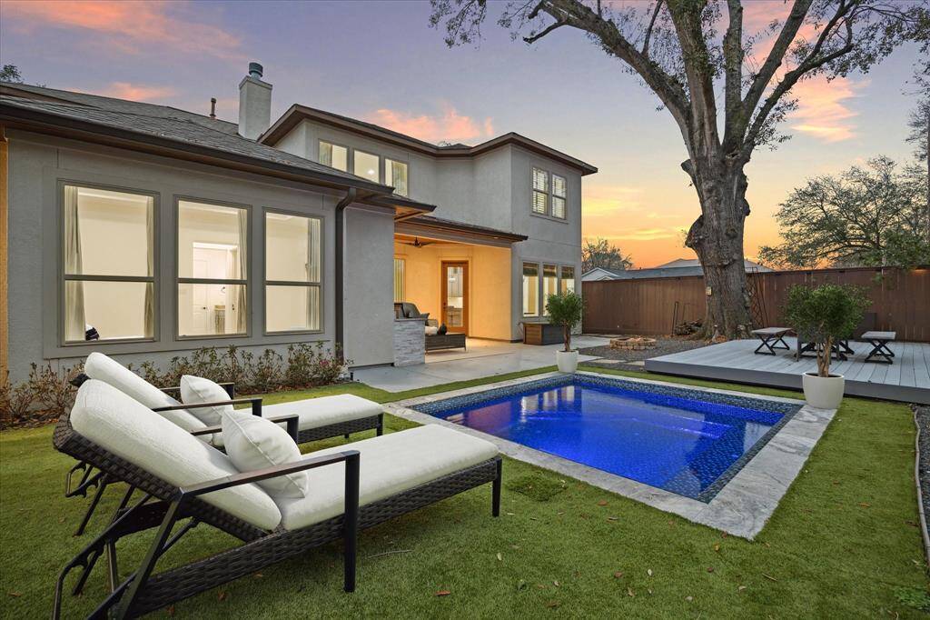 This backyard features a modern pool with surrounding greenery, a cozy patio area with seating, and ambient lighting. The contemporary home exterior complements the tranquil outdoor space, ideal for relaxation and entertaining.