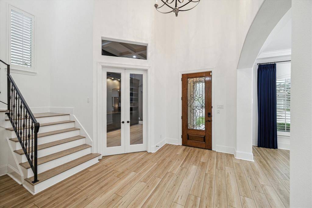 This entryway features a high ceiling and wood flooring, creating an inviting atmosphere. It includes a stylish wooden front door with decorative glass, a staircase with wrought iron railing, and double glass doors leading to another room. Natural light flows in through windows with shutters, enhancing the bright and open feel.